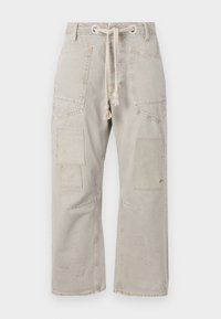 Béžové pracovní kalhoty s volným střihem, stahovacími šňůrkami v pase a několika kapsami; texturovaná tkanina s mírným opotřebením a drobnými skvrnami.