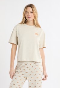 Beiges Baumwoll-T-Shirt mit kurzen Ärmeln und rundem Ausschnitt, mit einem kleinen Hundemotiv auf der linken Brust. Kombiniert mit beigen Pyjamahosen.