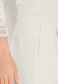 Close-up van een hand naast witte gestructureerde broeken met een riem en splitzakken, gecombineerd met een lange mouw witte top met kanten manchet.