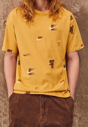 Żółta bawełniana koszulka z różnymi logo Nike i brązowym napisem "Just Do It". Krótkie rękawy, okrągły dekolt, luźny krój.