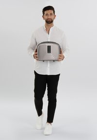 Een zilveren, hard-shell reiskoffer met ribbelstructuur en een rechthoekige vorm, vastgehouden door een persoon in een wit shirt en zwarte broek.