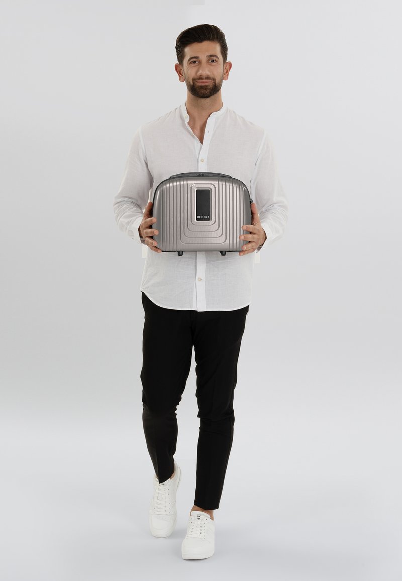 Een zilveren, hard-shell reiskoffer met ribbelstructuur en een rechthoekige vorm, vastgehouden door een persoon in een wit shirt en zwarte broek.