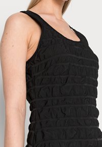 Schwarzes Tanktop mit einem strukturierten Design, vertikalen Rüschen und breiten Trägern. Der Stoff hat ein weiches, leicht zerknittertes Aussehen.