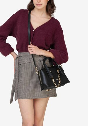Borsa nera con dettagli in catena dorata, forma rettangolare, texture liscia. Indossata con un maglione bordeaux e una mini gonna a quadri.