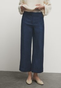 Granatowe spodnie o szerokich nogawkach wykonane z teksturowanej tkaniny, z wysokim stanem i podwiniętymi nogawkami, zestawione z jasnobeżowymi butami i kremowym swetrem.