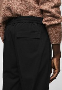 Osoba v hnědém texturovaném svetru a černých kalhotách s zadními kapsami, ukazuje část paže a oblasti pasu.