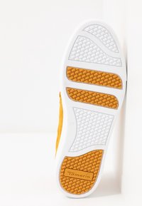 Semelle d'une chaussure jaune et blanche avec des sections de grip orange texturé et le nom de la marque « Tamaris » sur la zone du talon.