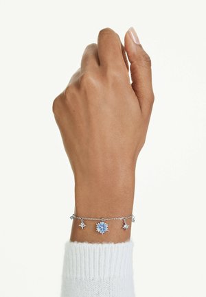 Mano con pelle marrone media che indossa un braccialetto d'argento con ciondoli a forma di stella con pietre preziose blu e trasparenti sopra un polsino bianco.