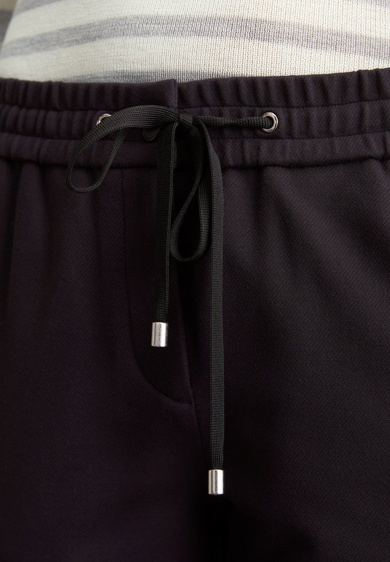 Schwarze Jogginghose mit einem Kordelzugbund, silbernen Akzenten an den Enden der Kordeln und einem strukturierten Stoff.