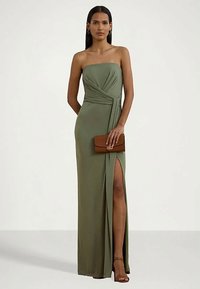 Kvinna i en ärmlös olivgrön klänning med sidospalt, som håller en brun kuvertväska, stående mot en helt vit bakgrund.