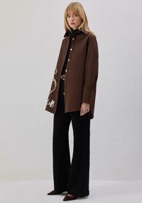 Pruun, üle suuruse nööpidega kinnitatav mantel tikandiga ühel küljel, millele on lisatud mustad laiade säärega püksid ja pruunid terava ninaga kingad.