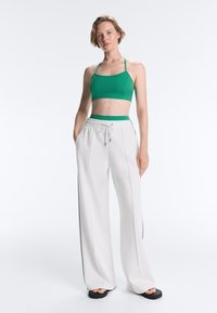 Femme portant un débardeur court vert et un pantalon blanc à jambes larges avec cordon, debout les mains dans les poches sur un fond blanc.