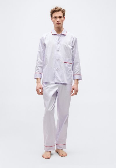 Νέος άνδρας που στέκεται ξυπόλητος, φορώντας λευκό σετ πυτζάμας με κόκκινη επένδυση, που αποτελείται από μακρυμάνικο πουκάμισο με κουμπιά και παντελόνι που ταιριάζει.
