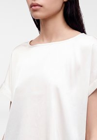 Jeune femme aux cheveux noirs et raides portant un haut blanc uni à manches courtes avec un col rond sur fond blanc.