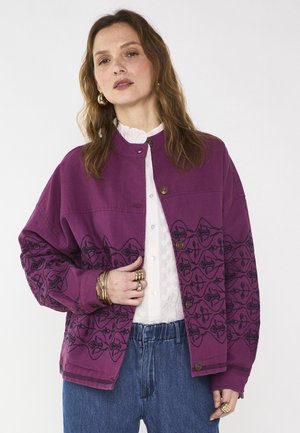 Vrouw draagt een paarse jas met knopen en zwarte bloemenpatronen, een wit blouse, blauwe spijkerbroek, gouden oorringen en meerdere gouden ringen en armbanden.