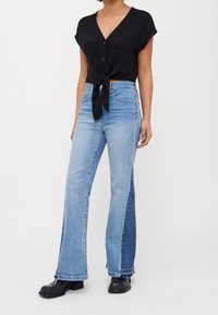 Donna che indossa un top nero a maniche corte con bottoni annodato in vita, jeans blu a due toni a campana e stivaletti neri chunky alla caviglia.