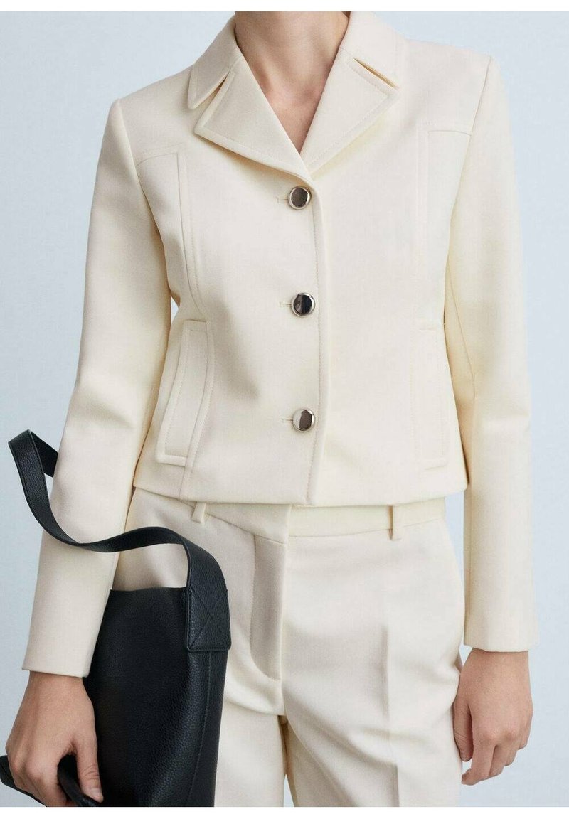 Femme portant une veste crème cintrée avec des boutons noirs et un pantalon assorti, tenant un sac à main en cuir noir par la bandoulière.