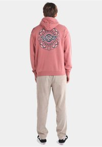 Sudadera rosa con un diseño gráfico circular en la parte posterior, combinada con pantalones de corduroy beige. Modelo de espaldas, usando zapatillas negras y blancas.