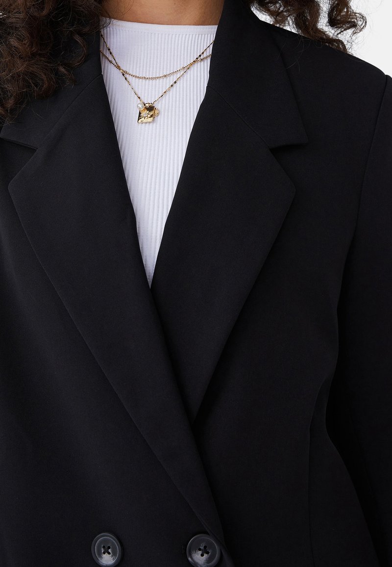 Kobieta w czarnym podwójnym żakiecie noszącym białą prążkowaną bluzkę i warstwowane złote naszyjniki, z których jeden ma kwadratowy wisiorek i czarę perłową.