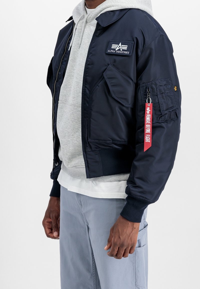 Homme portant une veste de vol Alpha Industries bleu marine avec une étiquette "Remove Before Flight" sur un sweat à capuche gris clair et un pantalon bleu clair.