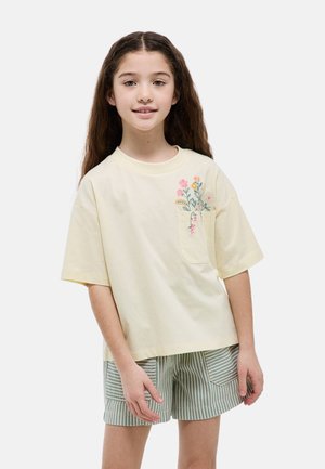 Fille aux longs cheveux bruns portant un T-shirt crème avec une broderie florale sur la poche et un short rayé vert, debout et souriante.