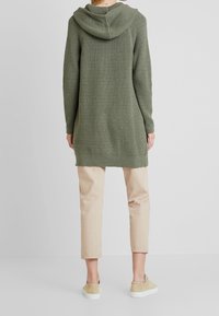 Olivenfarvet lang cardigan med hætte, struktureret strikkemønster og ribbet manchetter. Børnes over beigefarvede cropped bukser og beigefarvede slip-on sko.