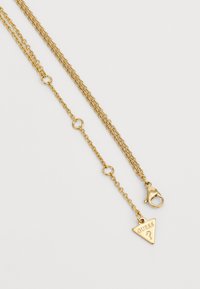 Collier en métal doré avec une conception allongée, un fermoir homard et un pendentif triangulaire portant l'inscription "GUESS".