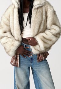 Veste en fausse fourrure crème avec capuche, présentant des manches larges et une texture douce sur un top court et une chemise à carreaux superposée en dessous.