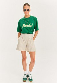 Zelené bavlnené tričko s nápisom "Merde!" v krémovom texte, skombinované s béžovými pruhovanými šortkami, zelenými teniskami a okrúhlymi slnečnými okuliarmi.