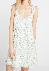 Bílé šaty se vzorem zelených listů, kulatým výstřihem a zavazovacími špagetovými ramínky. Lehce splývavá látka, elastický pas, krátká délka. Žádné výrazné kování ani akcenty.