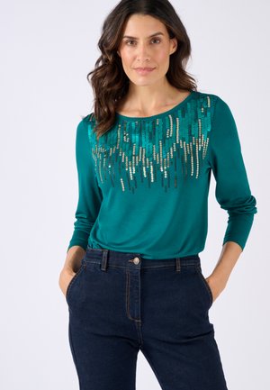 Top de manga larga en color aguamarina con lentejuelas decorativas en un patrón escalonado a lo largo del escote. Combinado con jeans de mezclilla oscura.