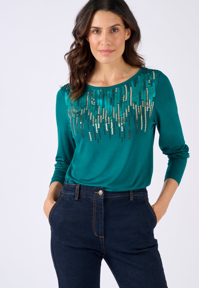 Teal langemouwtop met decoratieve pailletten in een verspringend patroon langs de halslijn. Gecombineerd met donkerdenim jeans.
