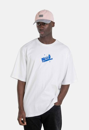 White short-sleeve t-shirt featuring blue graphic design, paired with a light pink cap with gray brim and logo patch. Black pants.