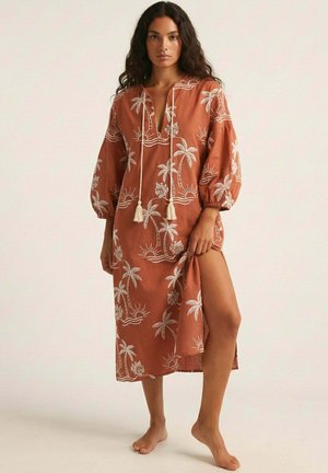 Mujer con cabello largo y rizado que lleva un vestido midi color óxido con estampados blancos de palmeras y sol, de pie descalza, sosteniendo la abertura del vestido.