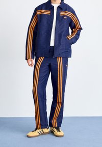 Mörkblå träningsoverall med orangea randiga detaljer, bestående av en mönstrad jacka och byxor, samt matchande skor. Dragkedja fram.