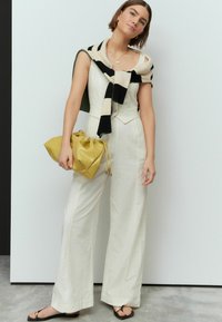 Combinaison beige clair à coupe ajustée, pull rayé noir et beige drapé sur les épaules, et sac à main avec cordon de serrage jaune.