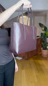 Bolso tote de cuero burdeos con patrón de diamante texturizado, asas largas y acabado suave, sostenido verticalmente sobre un suelo de madera.