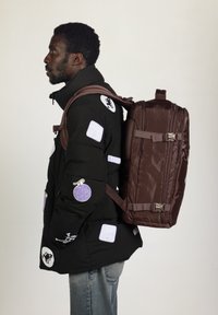 Doudoune noire avec divers patchs brodés, portée avec un sac à dos texturé marron foncé doté de sangles réglables et de détails en métal.