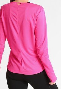 Osoba w jaskraworóżowej długorękawowej koszulce sportowej i czarnych legginsach, pokazana z tyłu na białym tle.