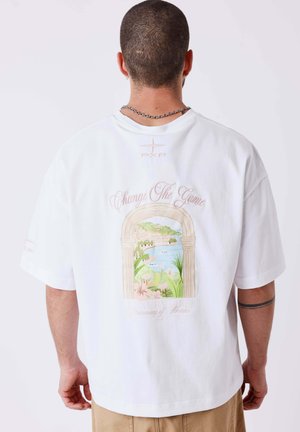 Man met kort haar en een ketting draagt een witte oversized T-shirt met de tekst "Change The Game" en een schilderachtig boogontwerp op de rug.