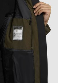 Wnętrze oliwkowozielonej kurtki z czarną podszewką, kieszenią na zamek i metką pielęgnacyjną. Miękka faktura z kontrastowymi materiałami i detalami w stichowaniu.