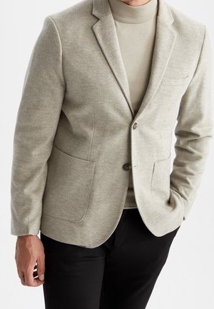 Homme portant un blazer beige texturé sur un col roulé beige clair, avec un pantalon noir, debout avec une main dans la poche.