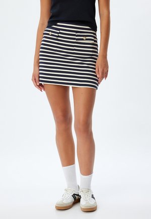 Falda a rayas en azul marino y crema, con dos bolsillos frontales y botones dorados. Llevada con calcetines deportivos blancos y zapatillas blancas.