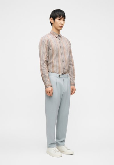 Άνδρας φορώντας πολύχρωμο ριγέ πουκάμισο με κουμπιά, ανοιχτόχρωμο μπλε παντελόνι και λευκά αθλητικά παπούτσια, στέκεται μπροστά σε λευκό φόντο.