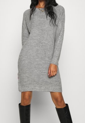 Kobieta ubrana w szarą dzianinową sukienkę z długimi rękawami i czarne wysokie kozaki za kolano, stojąca na białym tle.