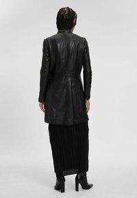 Cappotto lungo in pelle nera con vita aderente, maniche lunghe e una texture liscia. Indossato sopra una gonna nera a pieghe e stivali alla caviglia.