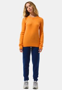 Maglione arancione realizzato in un tessuto morbido con maniche lunghe, abbinato a pantaloni della tuta blu scuro. L'outfit è completato da sneakers grigie.