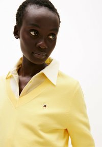 Geel v-hals sweater van zacht materiaal, gelaagd over een lichtgekleurde, geborduurde blouse. Bevat een klein, geborduurd logo op de borst.
