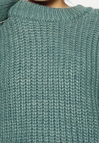 Chunky stickad tröja i mjuk teal, med ribbad textur och en hög, rundad halsringning. Materialet verkar varmt och mysigt.