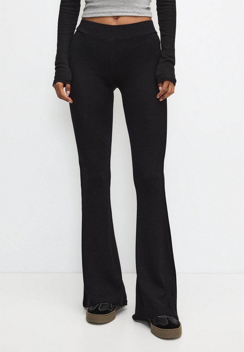 Pantalon noir évasé à côtes avec une taille haute. Le tissu a une texture douce, présentant une coupe ajustée qui s'évase à la cheville.